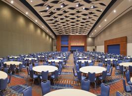 A brown and gray ballroom section with dozens of white 9 ft. round tables each surrounded by blue upholstered banquet chairs.