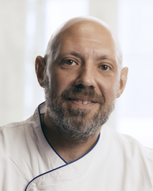 Chef Phil, a white man with a bald head and brown beard and mustache smiles at the camera in his kitchen uniform.