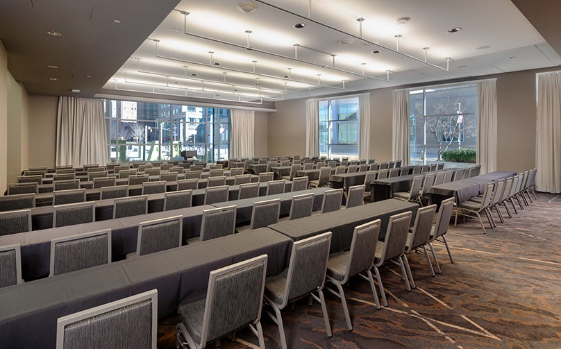 From the back of a meeting room showing open windows and filled with rows of rectangular tables and chairs and a podium in front.