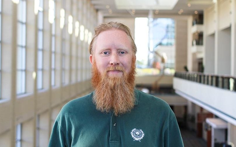 Headshot of Andrew Crane wearing a green polo standing on an indoor balcony overlooking large windows and a sculpture in background.