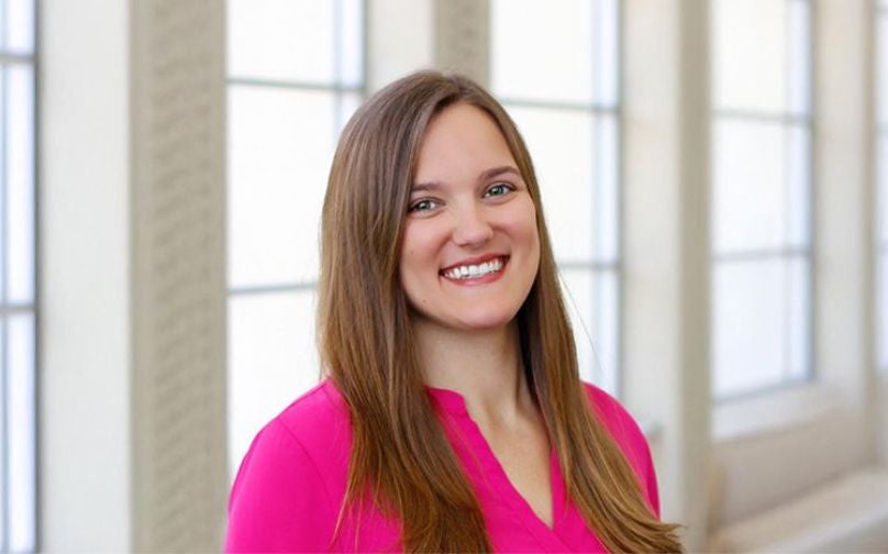Headshot of Ashley Auman wearing a pink top standing on an indoor balcony overlooking large windows.