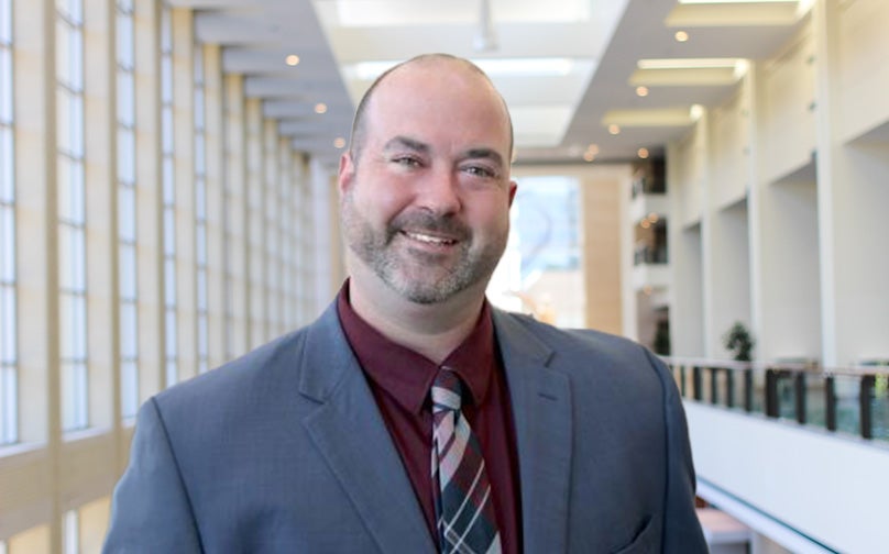 Headshot of Brad Clinton wearing a gray blazer, maroon shirt, and striped tie standing on an indoor balcony overlooking large windows and a sculpture in background.