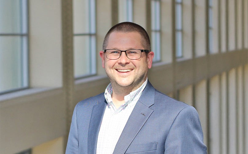 Headshot of Brian Clark wearing a gray blazer and white shirt standing on an indoor balcony overlooking large windows.