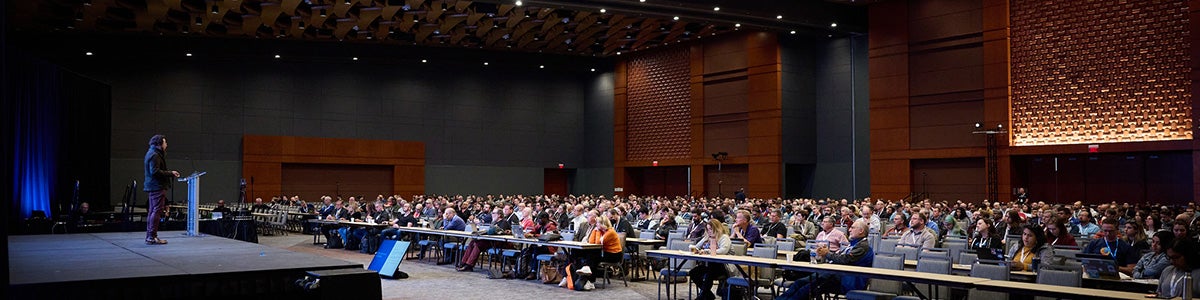 Ballroom with warm wood accents and dimmed lighting with a stage and speaker standing and audience listening while seated in a classroom setting.