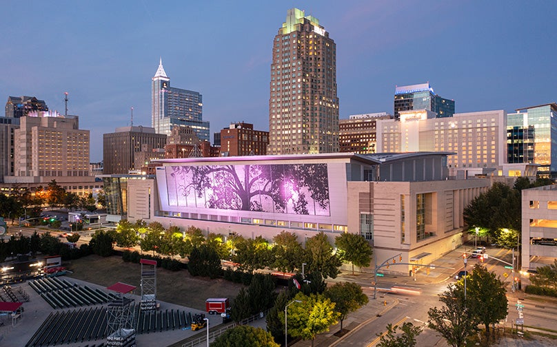 The Downtown Raleigh skyline at dusk, with the convention center and its illuminated shimmer wall featured prominently in the center.