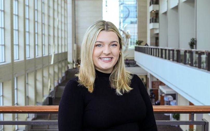 Headshot of Ellie King wearing a black turtleneck standing on an indoor balcony overlooking large windows and the convention center mezzanine.