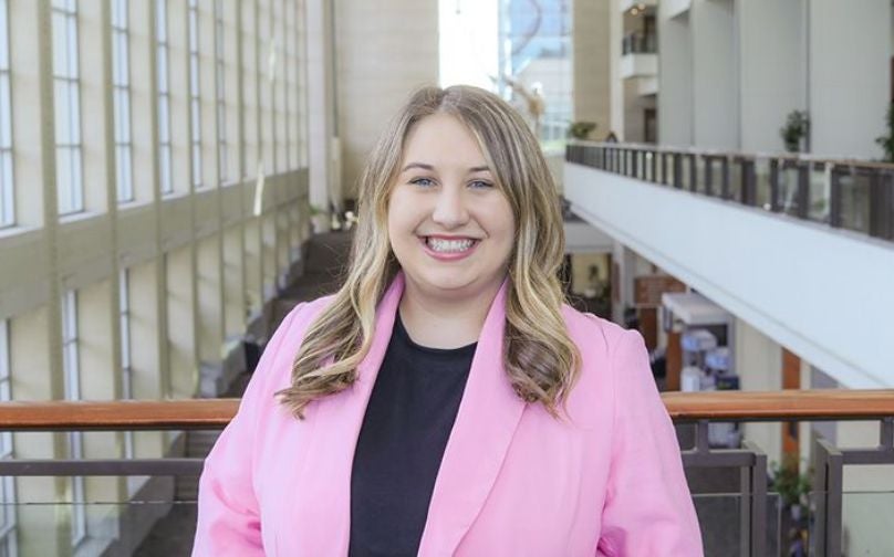 Headshot of Faith Dickson wearing a black top and pink blazer standing on an indoor balcony overlooking large windows and a the convention center mezzanine.