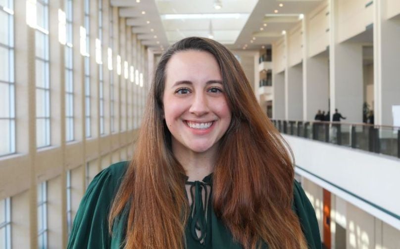 Headshot of Christina Alevizatos wearing a green top standing on an indoor balcony overlooking large windows.