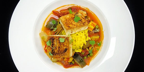 Close-up, birds eye view of white circular plate with seared white fish, yellow rice, vegetables, and orange sauce, lightly garnished with greens.