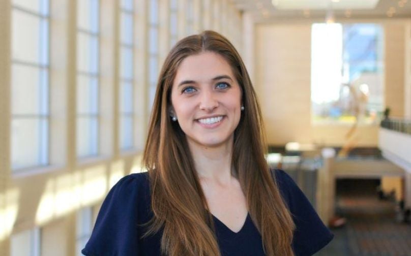 Headshot of Kate Jansen wearing a navy blue top standing on an indoor balcony overlooking large windows and a sculpture in background.