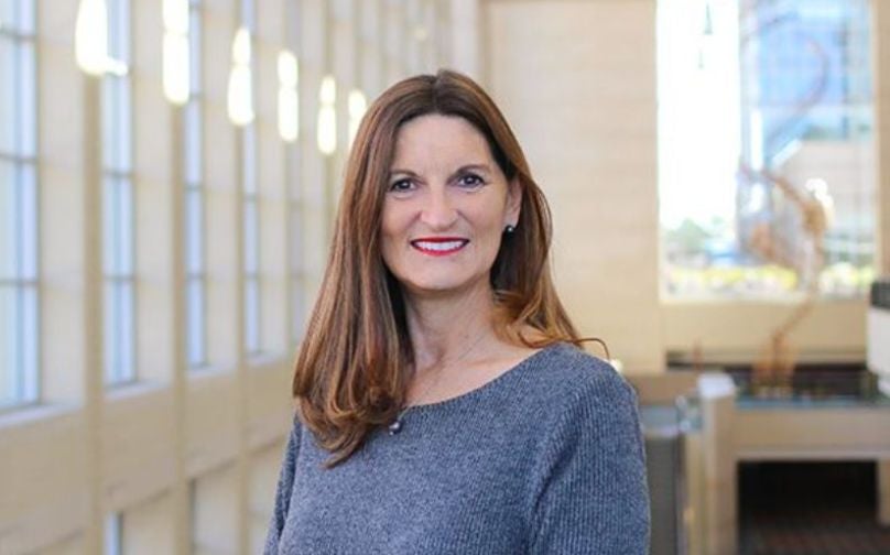 Headshot of Kerry Painter wearing a gray top standing on an indoor balcony overlooking large windows and a sculpture in background.