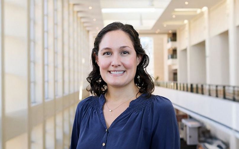 Headshot of Laura Knott wearing a blue top standing on an indoor balcony overlooking large windows and the convention center mezzanine..