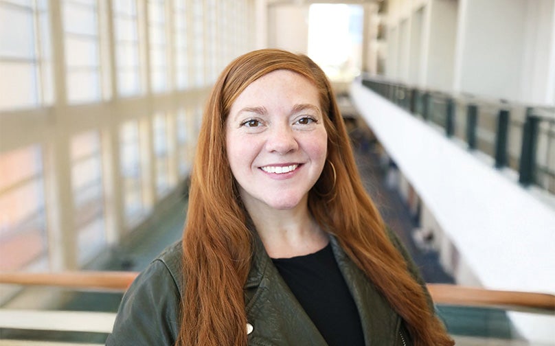 Headshot of Lauren Miller wearing a black top and leather jacket standing on an indoor balcony overlooking large windows and the convention center mezzanine.