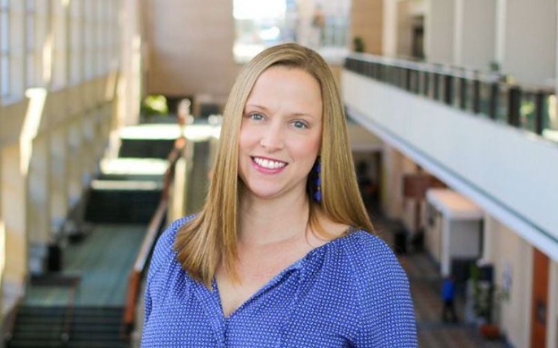 Headshot of Mara Craft wearing a blue top standing on an indoor balcony overlooking the mezzanine of the convention center.