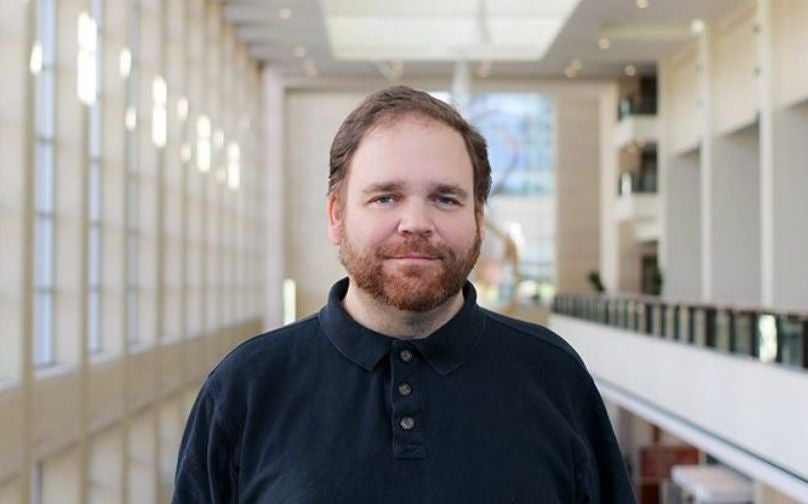 Headshot of Mike Dreese wearing a black polo standing on an indoor balcony overlooking large windows and a sculpture in background.