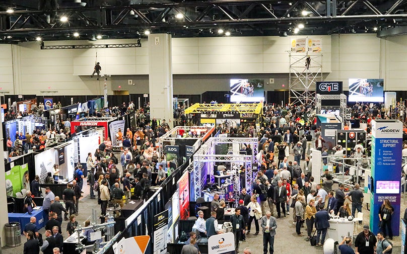 Trade show in an exhibit hall with vendor booths, attendees, and two men hanging from ceiling rafters in the back of the room.