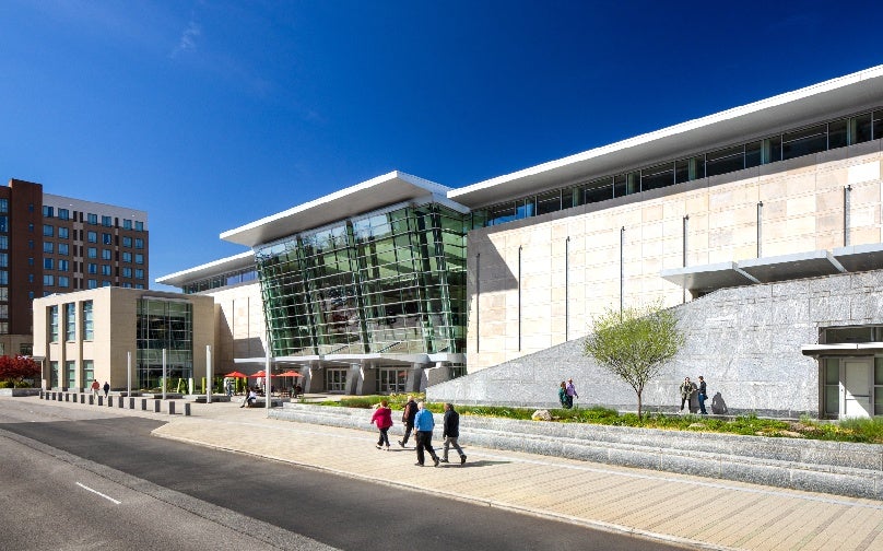 View of the entrance to Raleigh Convention Center from the side with people gathered and sitting at tables with red umbrellas.