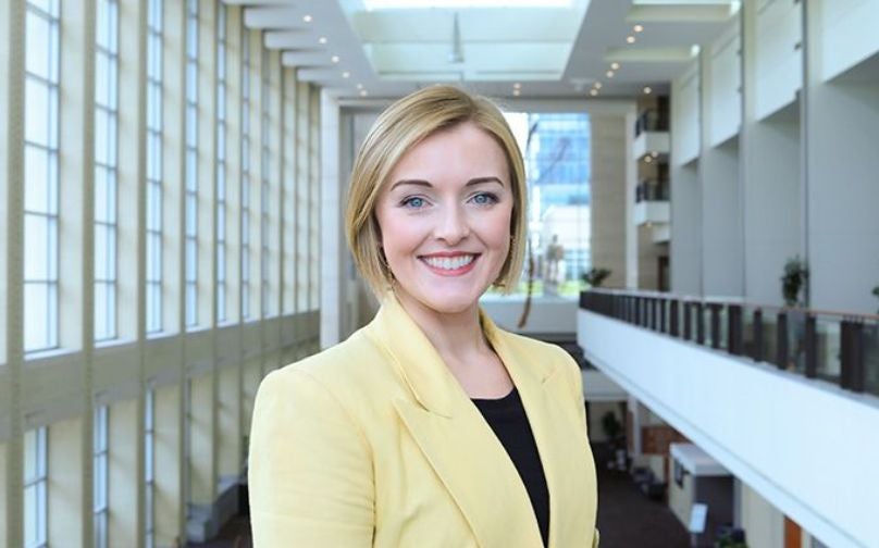Headshot of Olivia Holbrook wearing a black top and yellow blazer standing on an indoor balcony overlooking large windows and a sculpture in background.