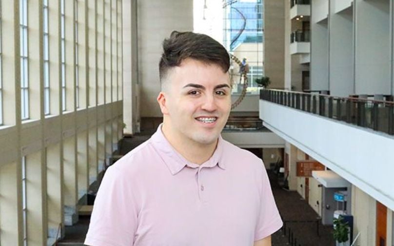 Headshot of Alex Bryant wearing a light pink polo standing on an indoor balcony overlooking large windows and a sculpture in background.