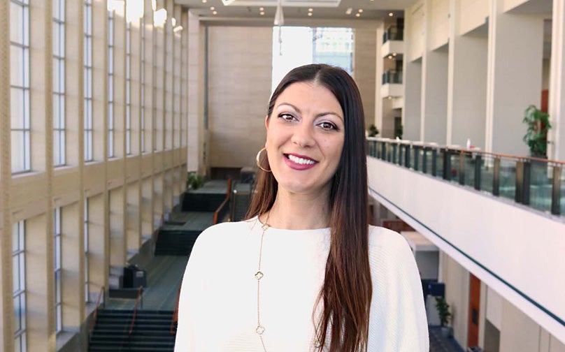 Headshot of Samantha Amodio wearing a white top standing on an indoor balcony overlooking large windows and the convention center mezzanine..
