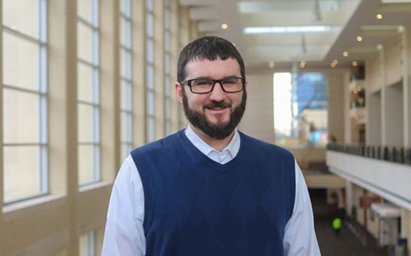 Headshot of Zachary Reed wearing glasses, a light blue long sleeve shirt with dark blue vest, standing on an indoor balcony overlooking large windows and a sculpture in background.