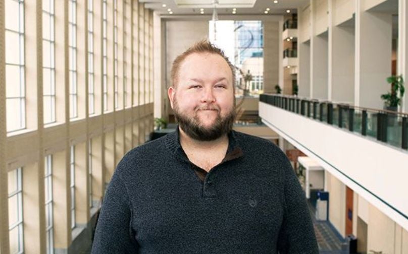 Headshot of Zenik Hite wearing a black polo standing on an indoor balcony overlooking large windows and a sculpture in background.