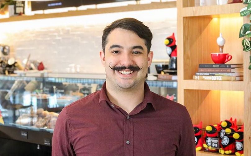 Headshot of Anthony wearing a burgundy button up standing in front of the baked goods counter and shelves with books and cardinal plushies.