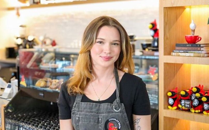 Headshot of Jess wearing a black polo and Complex Brew branded apron standing in front of the baked goods counter and shelves with books and cardinal plushies.