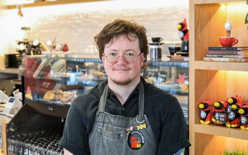 Headshot of Mael wearing a black polo and Complex Brew branded apron standing in front of the baked goods counter and shelves with books and cardinal plushies.