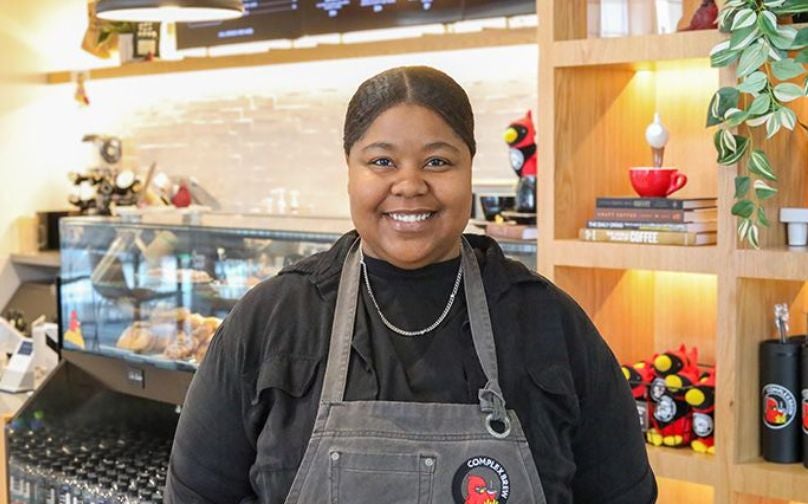 Headshot of Paige wearing a black polo and Complex Brew branded apron standing in front of the baked goods counter and shelves with books and cardinal plushies.