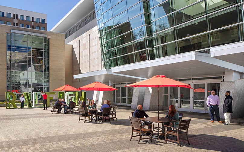 View of the entrance to Raleigh Convention Center from the side with people gathered and sitting at tables with red umbrellas..