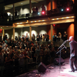 Photo taken from the stage, where on the left you can see a full audience, standing and clapping. On the right, the performer standing on the stage in front of a mic.
