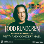 Todd Rundgren staring into the distance with a colorful psychedelic background with radiating light patterns in shades of green, orange, pink, blue, and purple, creating a vibrant, swirling effect.