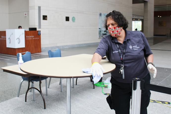 A white female RCC staff member with short, black wavy hair wearing a gray RCC uniform polo shirt and dark-colored pants wipes down tables in the Main Lobby with a Security Stand in the background.
