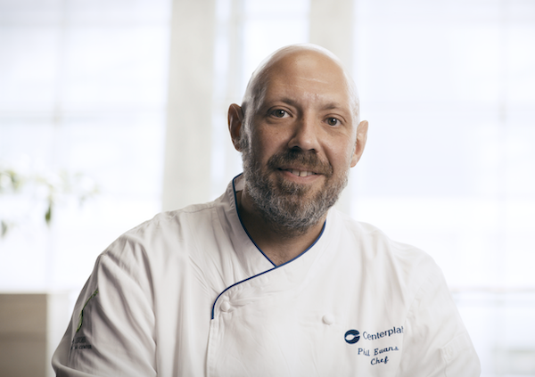 A middle-aged white man with a shaved head and beard wearing his kitchen whites (a white branded uniform) smiling into the camera.