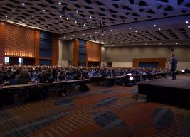 Ballroom full of people seated facing a speaker on stage 