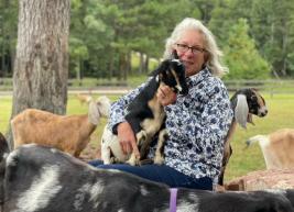 A seated white woman with shoulder-length white hair and glasses in a patterned white and blue floral long-sleeved jacket holding a small black, brown, and white goat. There are other goats standing near her and she is in a green field with grass and trees.