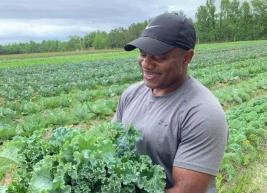 A smiling middle-aged muscular Black man wearing a gray tee-shirt and black baseball cap holding and looking at leafy greens while standing in a big green field below an overcast sky.