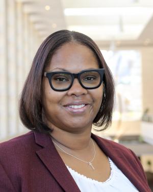 A black female with short medium brown hair is wearing black-rimmed glasses, a white top and a maroon blazer with the sun-filled RCC mezzanine in the background.