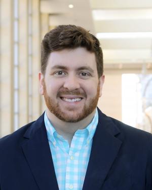 A white man with brown hair and facial hair smiles at the camera. He is wearing a green and white gingham button-down shirt with a navy blue blazer.