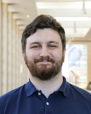 A white man with short brown hair and facial hair smiles at the camera. He is wearing a navy blue polo shirt.