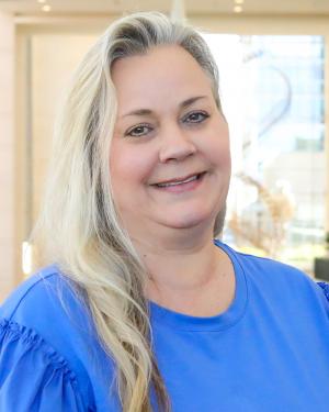 Blonde-white haired white woman smiles at the camera. She has brown eyes and her long hair is pulled to one side. She wears a blue long-sleeved top.