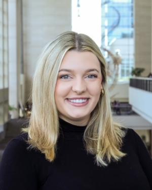 A smiling blonde woman looks into the camera. She has blue-green eyes and is wearing a black turtleneck. 