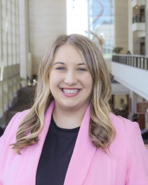 Smiling woman with blonde hair looking into the camera, wearing a pink blazer and black shirt. She is standing inside, in front of bright windows.