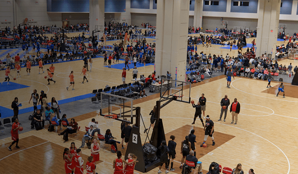Beige basketball courts set up in the Exhibit Hall for the Deep South Classic basketball tournament with spectators sitting along the sidelines and teenaged female players on the court playing in a game.